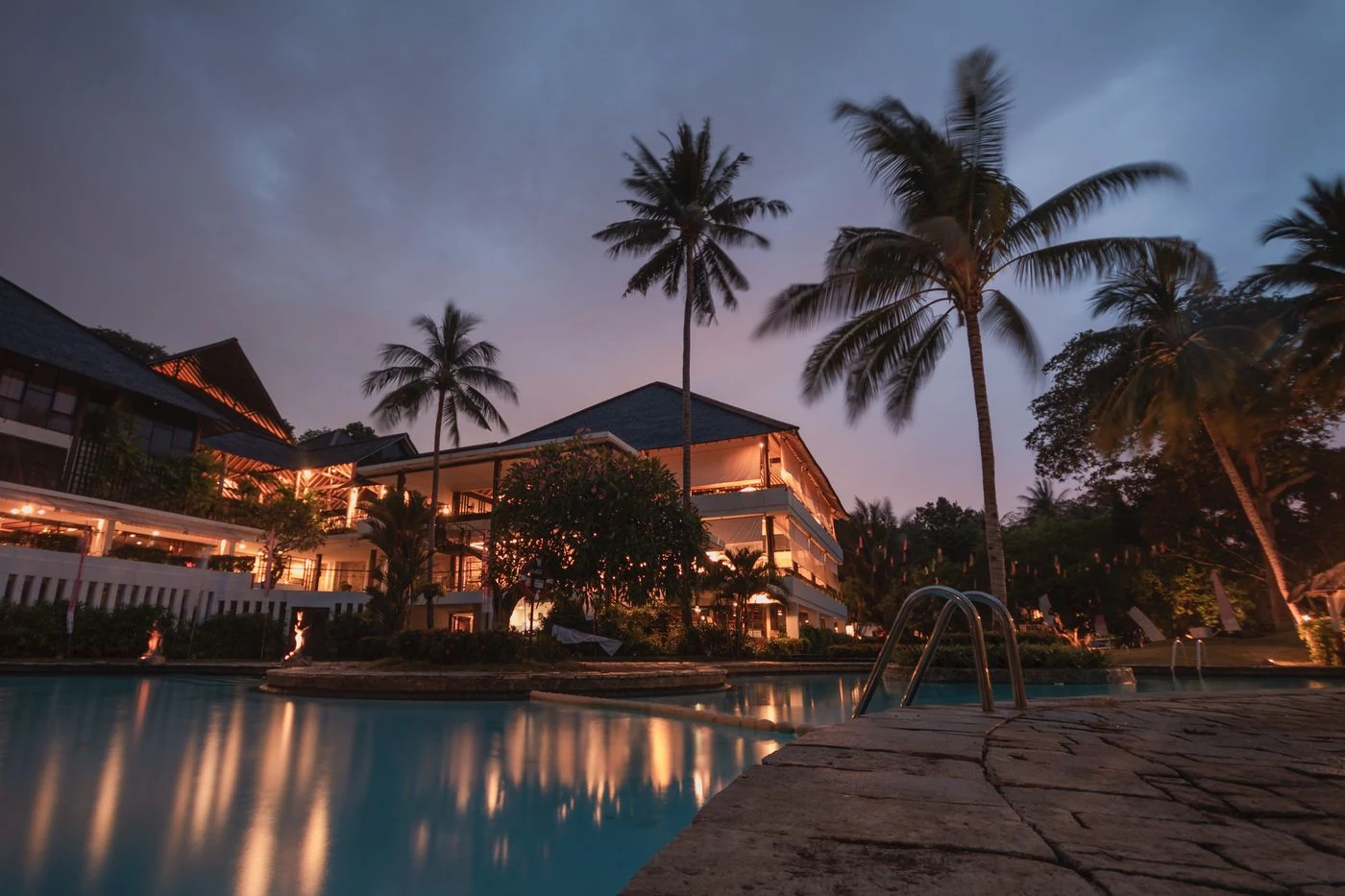 Palm-lined hotel swimming pool with warmly lit buildings at dusk